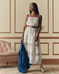 A woman stands indoors beside a pink upholstered chair and paneled wall, wearing THE GREAT. SP26 D2's The Rosette Crochet Dress in cream and holding a blue jacket.