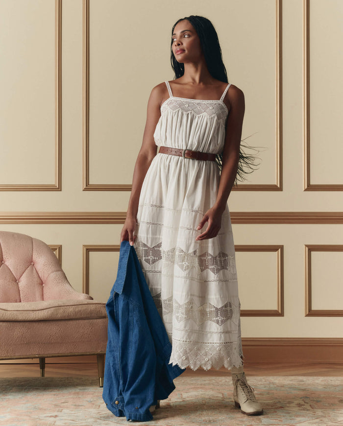 A woman stands indoors beside a pink upholstered chair and paneled wall, wearing THE GREAT. SP26 D2's The Rosette Crochet Dress in cream and holding a blue jacket.