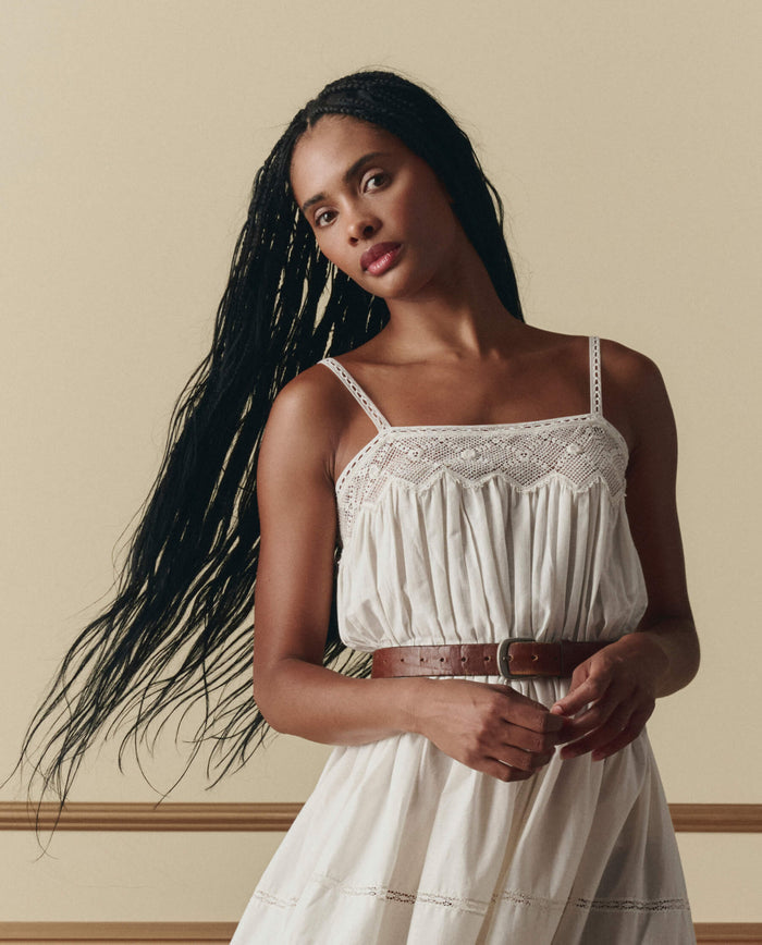 A woman with long braided hair wears THE GREAT.'s The Rosette Crochet Dress in cream, standing against a beige background.