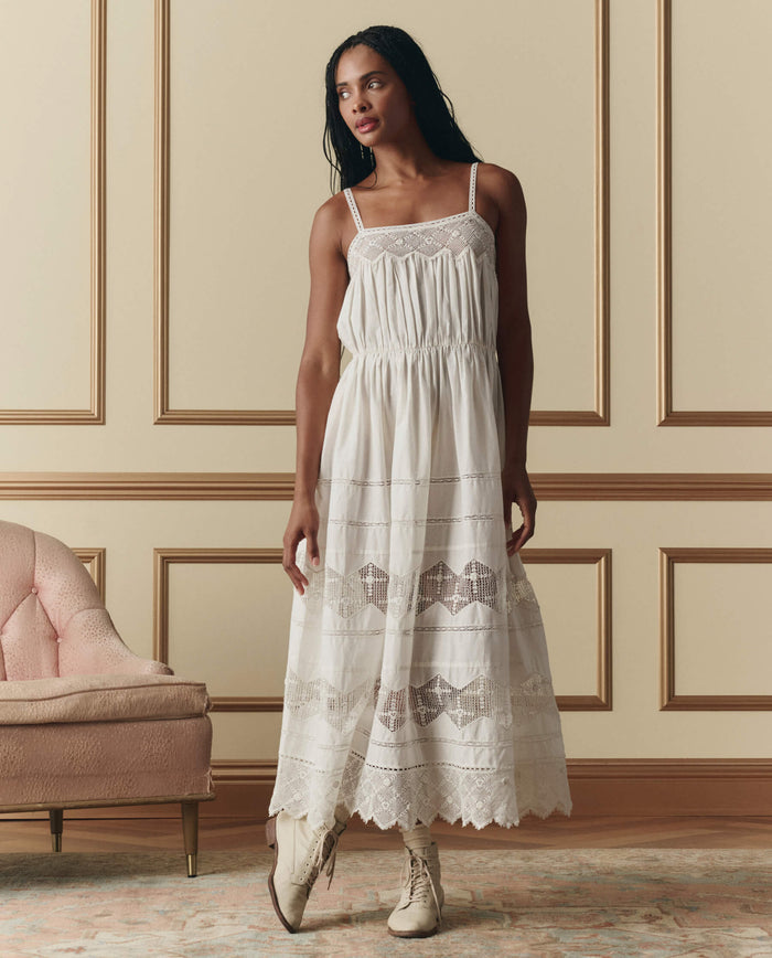 A woman stands indoors next to a pink chair and paneled wall, wearing THE GREAT. SP26 D2 The Rosette Crochet Dress in cream with lace details and beige ankle boots.