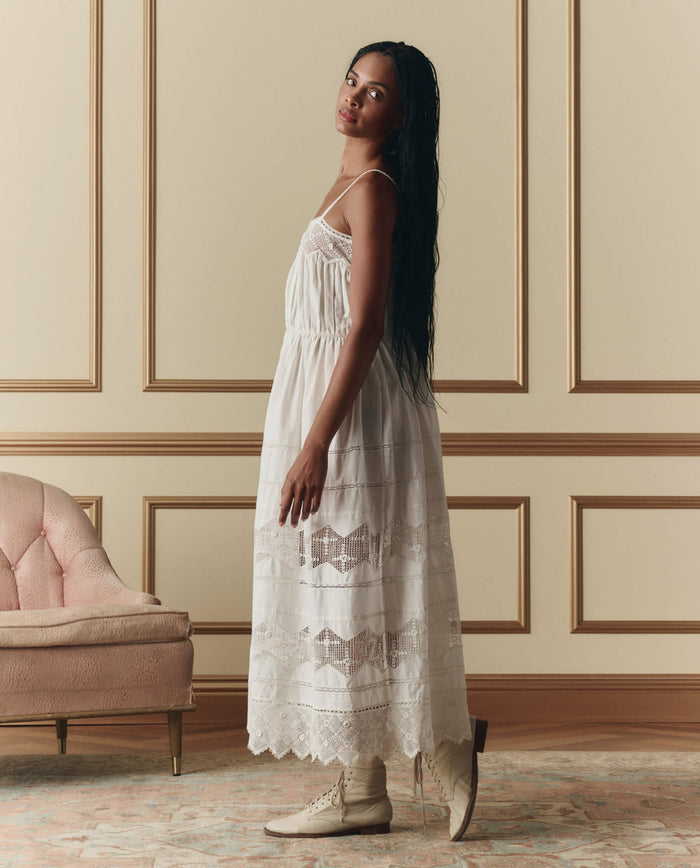Woman wears THE GREAT. SP26 D2 The Rosette Crochet Dress in cream, paired with cream boots, standing sideways in an elegant room with beige paneled walls, a pink velvet sofa, and a patterned rug.