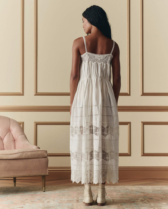 A woman stands with her back to the camera, wearing THE GREAT. SP26 D2 The Rosette Crochet Dress in cream and cream boots, in a room with beige paneled walls and a pink upholstered chair.