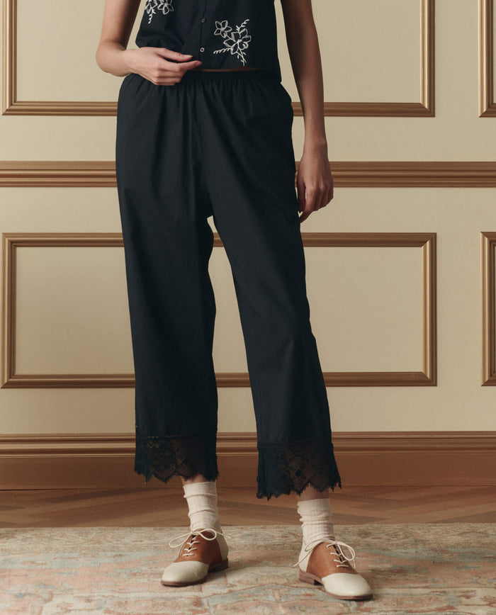 A person stands indoors wearing The Rosette Crochet Pant in black by THE GREAT. SP26 D2, paired with a black top featuring white embroidery, cream socks, and two-tone oxford shoes.