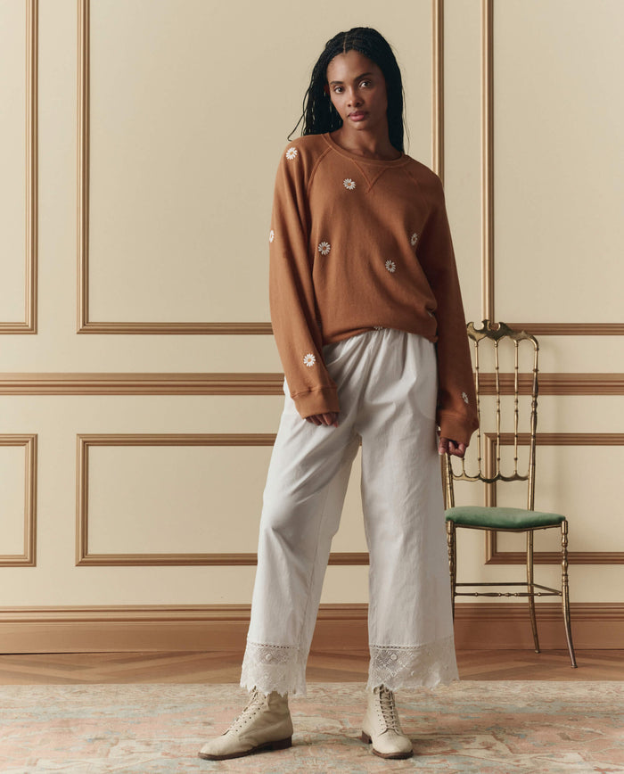 Indoors, a woman wears THE GREAT. SP26 D2’s The Rosette Crochet Pant in cream, paired with a brown floral sweater and beige boots; a green chair sits in the background.