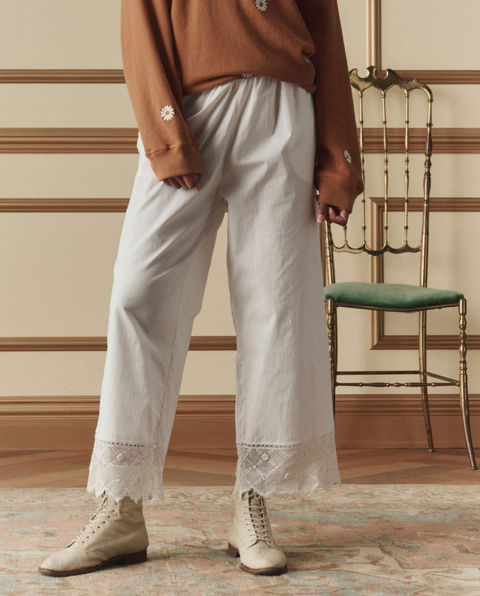 A person wearing a brown long-sleeve shirt, THE GREAT. SP26 D2 The Rosette Crochet Pant in cream, and beige lace-up boots stands indoors near a gold chair with a green cushion.