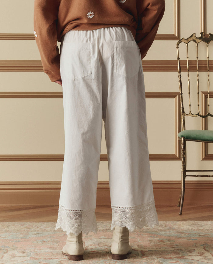 A person stands indoors wearing THE GREAT. SP26 D2 The Rosette Crochet Pant in cream, styled with cream boots and a brown sweater, beside a wooden chair and decorative wall paneling.