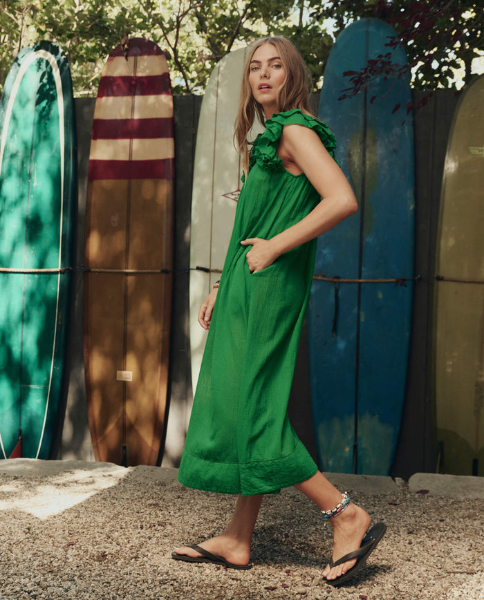 A woman in THE GREAT. SP26 SWIM The Ruffle Coverup – Bright Sprig stands outdoors on gravel, colorful surfboards lined up behind her.
