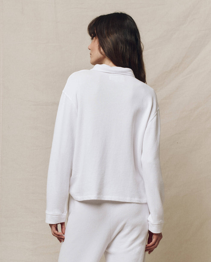 A woman wearing "The Rugby Sweatshirt" in True White from THE GREAT. SP25 POLO LOUNGE stands facing away on a beige background, resembling casual sportswear.