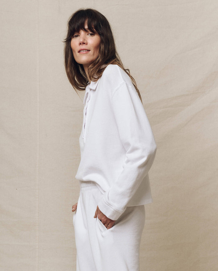 A person in THE GREAT. SP25 POLO LOUNGE's "The Rugby Sweatshirt" in True White, paired with pants, captures the essence of casual wear against a beige backdrop, gazing slightly at the camera.