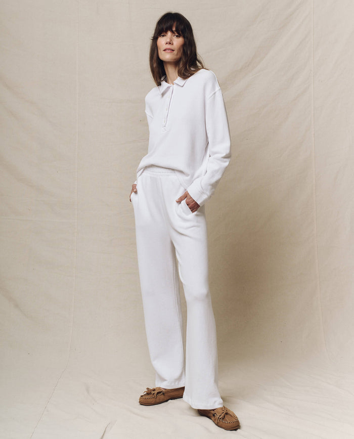 A person in The Rugby Sweatshirt by THE GREAT. SP25 POLO LOUNGE, paired with matching loose pants and brown shoes, stands with hands in pockets on a neutral fabric backdrop, effortlessly showcasing casual wear with sporty elegance.