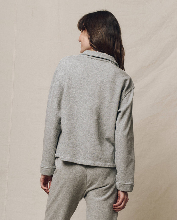 Against a neutral fabric backdrop, a person with long hair is seen from behind wearing THE GREAT. SP25 POLO LOUNGE's The Rugby Sweatshirt in Varsity Grey, paired with pants, exuding casual comfort.