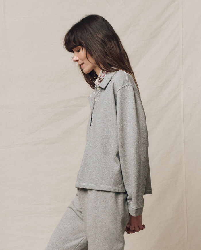 A person with long brown hair is wearing THE GREAT. SP25 POLO LOUNGE's The Rugby Sweatshirt in Varsity Grey with matching pants, standing against a beige backdrop.