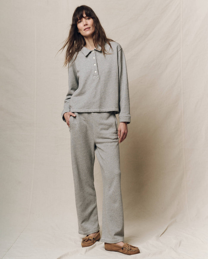 On a beige backdrop, a person wears THE GREAT. SP25 POLO LOUNGE's "The Rugby Sweatshirt" in Varsity Grey with matching pants, hands in pockets, and brown moccasin slippers.