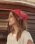 A woman in a white top wears THE GREAT. SU26's The Sailboat Bandana in red, standing in profile before a wooden window frame and beige siding.