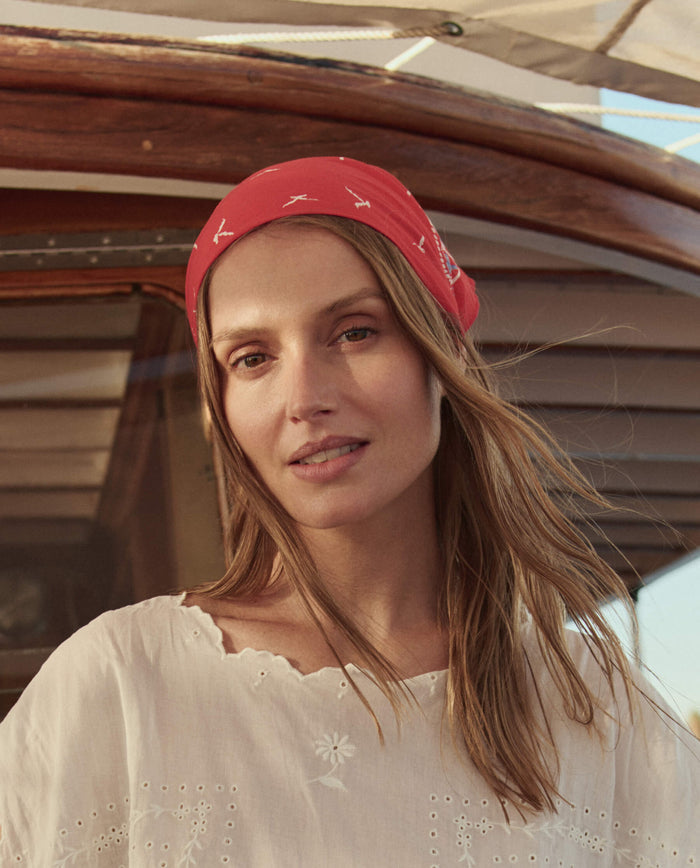 A woman with long hair wears THE GREAT. SU26's The Sailboat Bandana in Red and a white blouse, standing before a wooden boat structure.