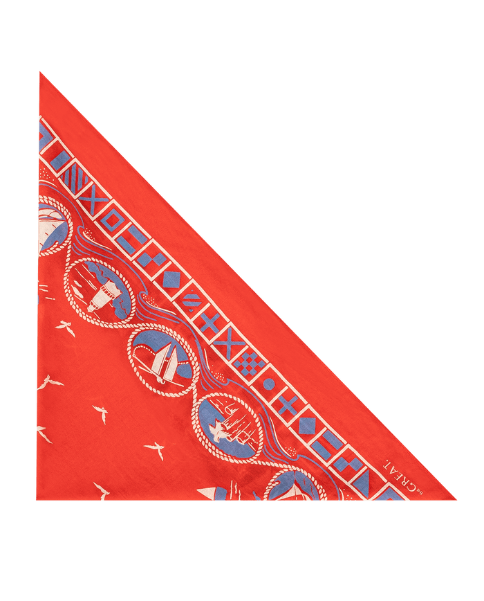 The Sailboat Bandana by THE GREAT. SU26—red silk, folded into a triangle, with blue and white nautical designs, small bird motifs, and "THE GREAT. SU26" in the corner—makes an elegant sailing accessory.