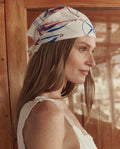 A woman with long hair wears THE GREAT. SU26 The Sailboat Bandana in white, styled on her head, paired with a white sleeveless top and standing indoors by a wooden surface.