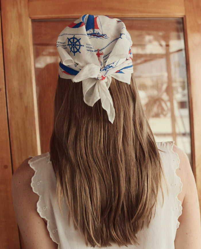 A woman with long light brown hair faces away, wearing THE GREAT. SU26 The Sailboat Bandana in White as a sailboat accessory and a white sleeveless top with eyelet trim.