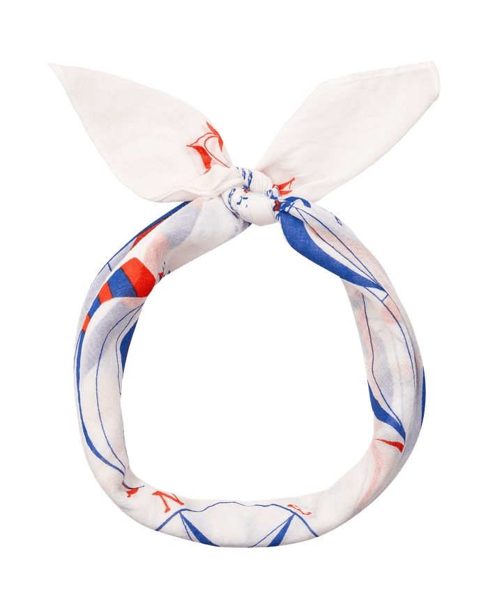 The Sailboat Bandana by THE GREAT. SU26 is a white nautical accessory featuring blue and red patterns, tied in a circle with two pointed ends at the top.