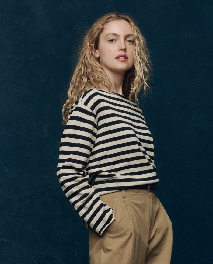 A woman with wavy blonde hair stands against a dark background, wearing THE GREAT. SP25 COLLECTORS "The Sailor Sweater" in Black Stripe and tan pants.