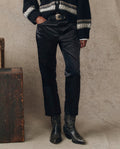 Wearing THE GREAT. H25 COLLECTION’s The Satin Wayne pants in black, paired with a black patterned sweater, a statement belt, and black cowboy boots, a person stands by a wooden box against a neutral backdrop.