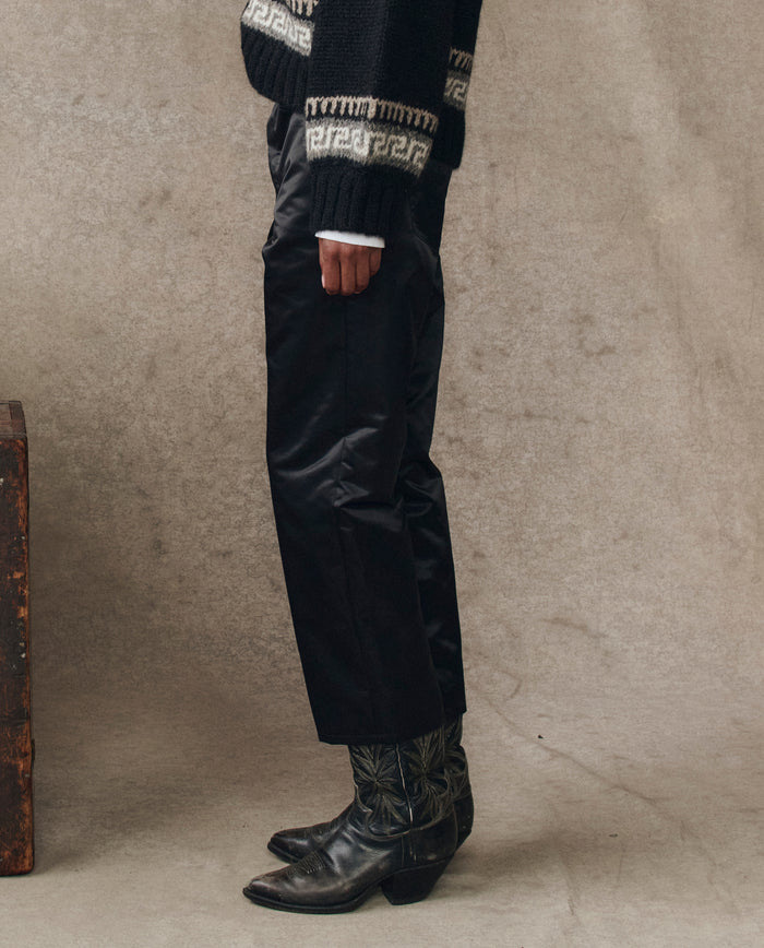 Wearing THE GREAT. H25 COLLECTION’s The Satin Wayne in black, this person pairs black pants, patterned dark boots, and a geometric-sleeved sweater to effortlessly showcase refined satin style.