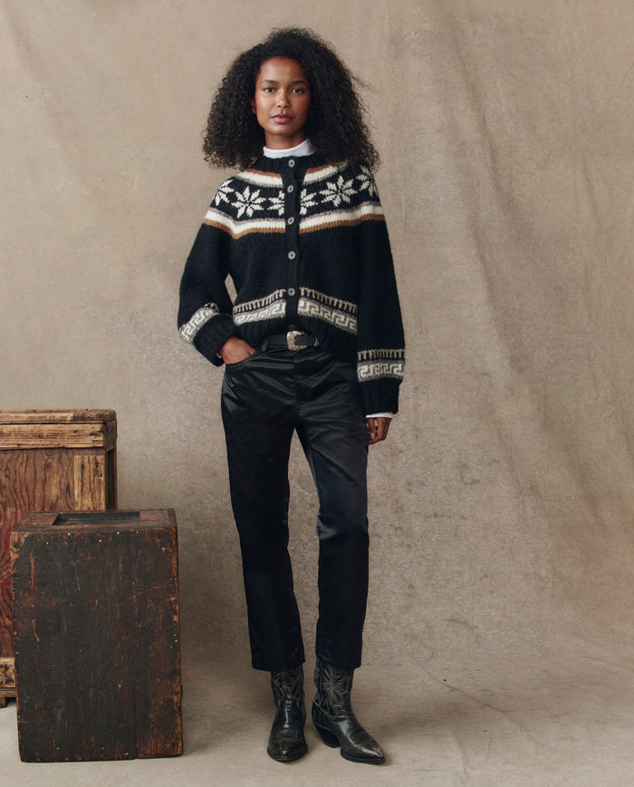 A woman poses before a beige backdrop, wearing THE GREAT. H25 COLLECTION's The Satin Wayne in black, paired with black pants, a belt, and black boots. Wooden boxes sit beside her, while satin accents elevate her look.