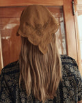 A person with long light brown hair wears THE GREAT. SU26 The Scalloped Bandana in Clay, paired with a dark patterned top, seen from behind indoors.