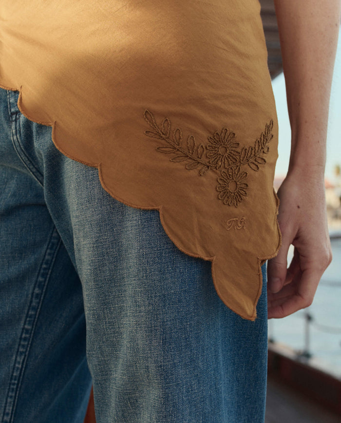 Outdoors, a person wears blue jeans, a mustard-yellow embroidered top with a scalloped hem, and THE GREAT. SU26 The Scalloped Bandana in Clay.