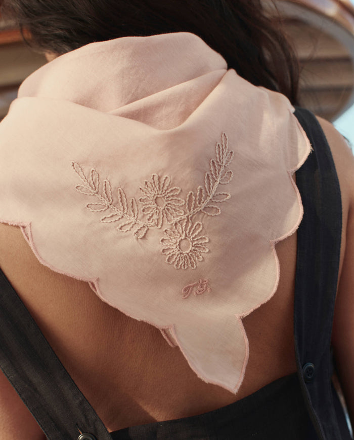 A person wears The Scalloped Bandana in Petal by THE GREAT. SU26—light pink, embroidered with a floral design and scalloped edges—over a dark sleeveless top.