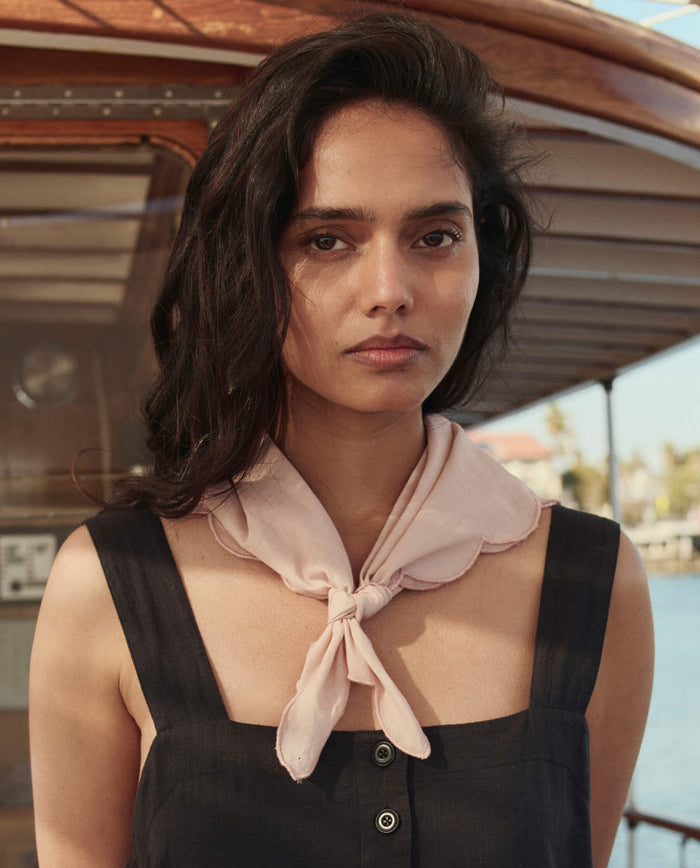 A woman with long dark hair, in a sleeveless black top, wears THE GREAT. SU26 The Scalloped Bandana in Petal around her neck while standing before a boat on a sunny day.
