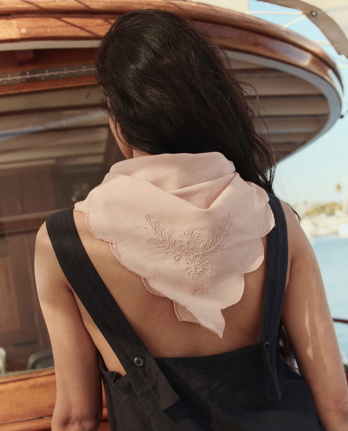A person with long dark hair faces away on a boat, wearing a sleeveless black outfit and THE GREAT. SU26’s The Scalloped Bandana in Petal, featuring light pink embroidery and draped over their shoulders.