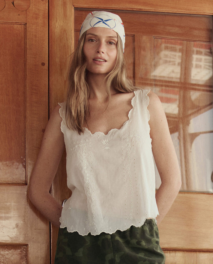 A woman wearing THE GREAT. SU26 The Eyelet Tank in white, green camouflage pants, and a patterned headscarf stands in front of a wooden door.