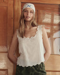 A woman wearing THE GREAT. SU26 The Eyelet Tank in white, green camouflage pants, and a patterned headscarf stands in front of a wooden door.