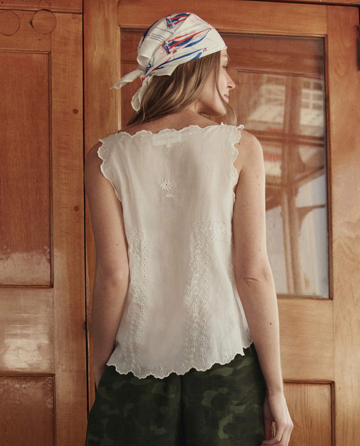 A woman stands with her back to the camera, wearing THE GREAT. SU26 The Eyelet Tank in white, camouflage shorts, and a patterned headscarf, in front of a wooden door.