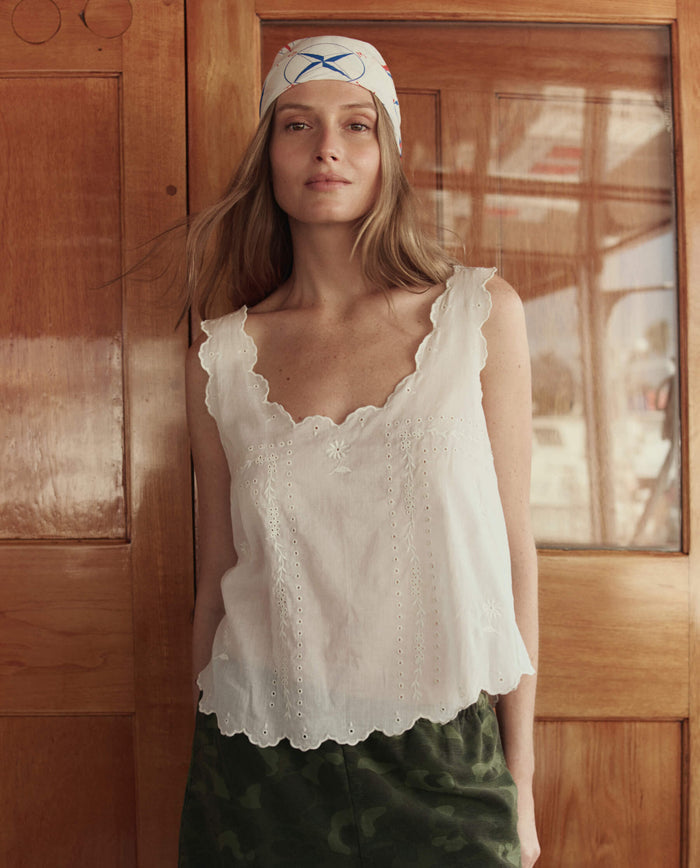 A woman wears THE GREAT. SU26 The Eyelet Tank in white, styled with a patterned headscarf, standing indoors before wooden doors and looking at the camera.