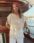 A woman wears THE GREAT. SU26 The Eyelet Tunic in white, standing on a wooden boat with water and docked boats in the background.