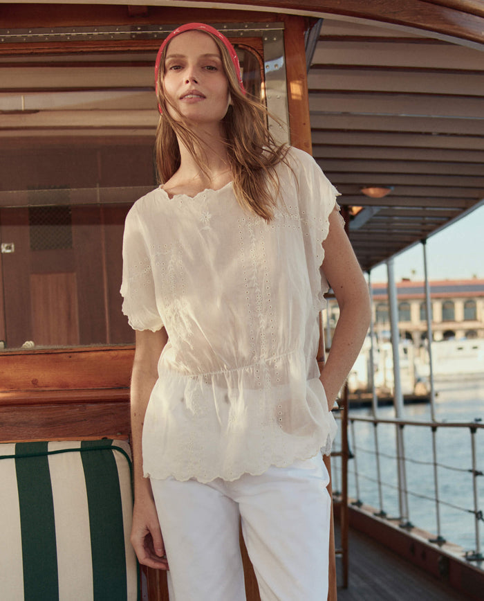 A woman in THE GREAT. SU26 The Eyelet Tunic in white and matching pants stands on a wooden boat, with water and buildings behind her.