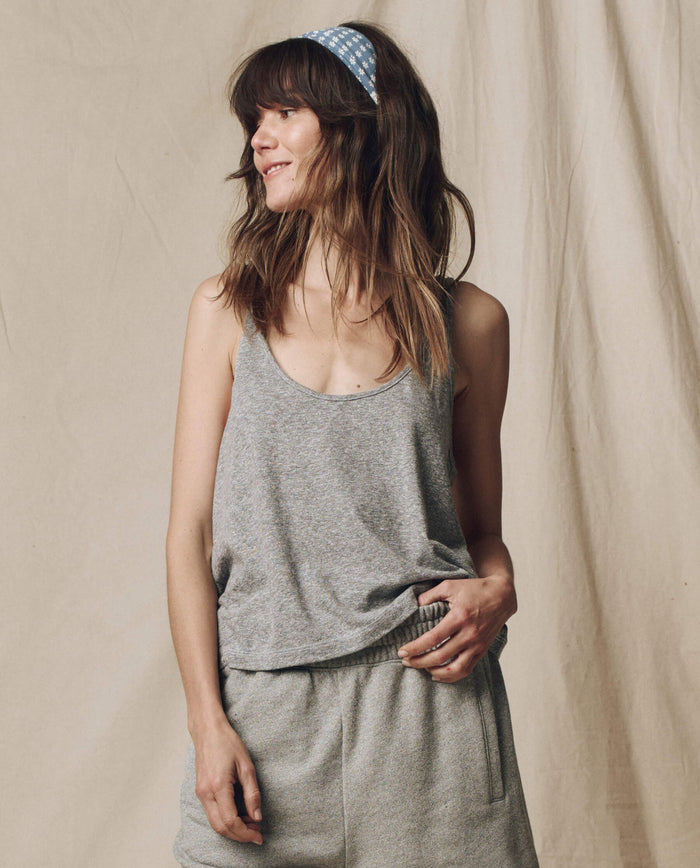 A woman with loose hair and a blue polka-dot headband smiles to the side, wearing THE GREAT. SU25 CORE KNITS' The Scoop Neck Tank in Heather Grey, styled with shorts against a beige fabric backdrop.