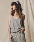 A woman with loose hair and a blue polka-dot headband smiles to the side, wearing THE GREAT. SU25 CORE KNITS' The Scoop Neck Tank in Heather Grey, styled with shorts against a beige fabric backdrop.