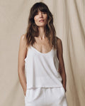 A woman with long brown hair stands against a neutral fabric backdrop wearing THE GREAT. SU25 CORE KNITS Scoop Neck Tank in True White and white pants with her hands in her pockets for a relaxed, effortless look.