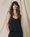 A woman with long brown hair wears THE GREAT. SU25 CORE KNITS The Scoop Neck Tank in Almost Black, standing before a beige fabric backdrop and looking at the camera with a neutral expression.