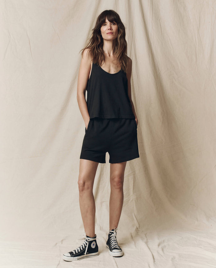 A woman stands before a beige fabric backdrop wearing THE GREAT. SU25 CORE KNITS The Scoop Neck Tank in Almost Black, paired with black shorts and black high-top sneakers, her hands tucked in her pockets.