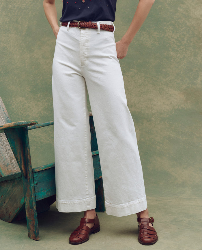 A person wears The Seafair Jean in True White by THE GREAT. SP25 D1, styled with a brown braided belt and woven leather shoes, standing beside a rustic wooden chair against a textured green backdrop.