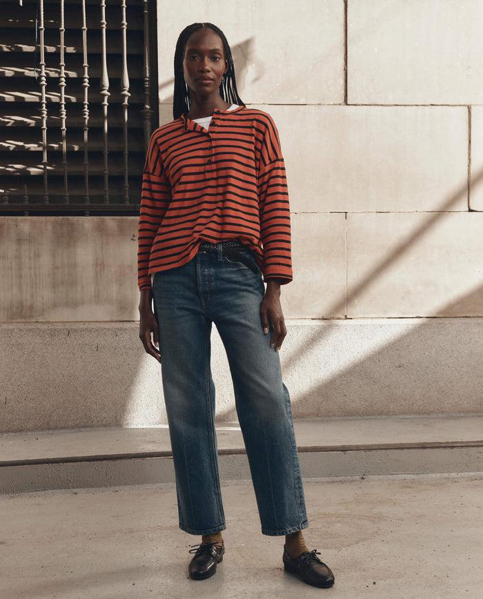 A person stands outdoors in front of a stone wall, wearing THE GREAT. F26 D1 The Shrunken Henley in Hot Red Naval Stripe, paired with blue jeans and black shoes, their expression neutral.