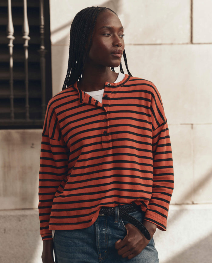 A person wearing THE GREAT. F26 D1 The Shrunken Henley in Hot Red Naval Stripe and jeans stands in front of a light-colored wall, looking to the side.