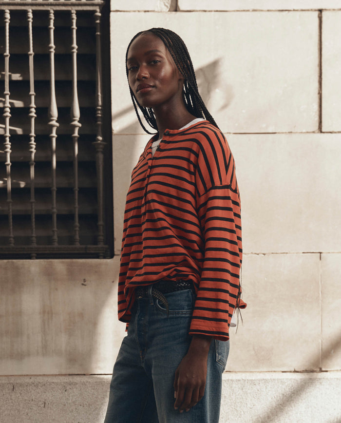 Wearing THE GREAT. F26 D1’s The Shrunken Henley in Hot Red Naval Stripe and jeans, a person stands outdoors near a beige wall with a black metal window grate.