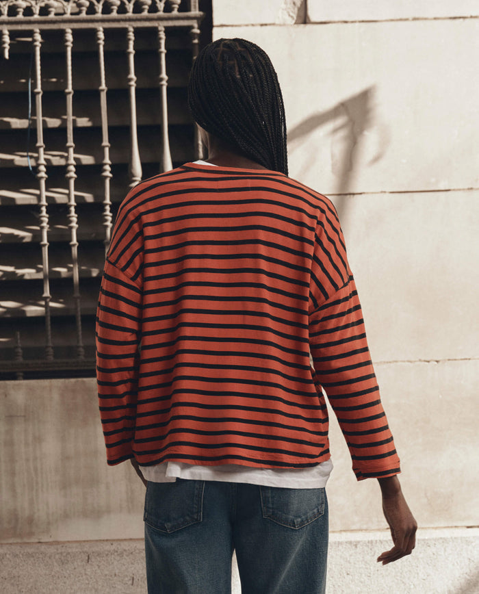 A person with long braided hair stands with their back to the camera, wearing THE GREAT. F26 D1 The Shrunken Henley in Hot Red Naval Stripe over a white tee and blue jeans, positioned in front of a metal grate.