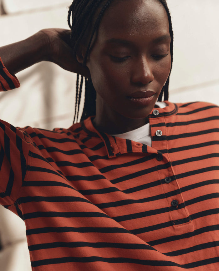 A person wearing THE GREAT. F26 D1 The Shrunken Henley in Hot Red Naval Stripe stands with one arm raised, looking downward.