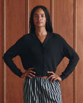 A person stands in front of a wooden panel wall, wearing THE GREAT. SP26 D1 The Shrunken V Neck Cardigan in black and striped pants, with hands on hips and a neutral expression.